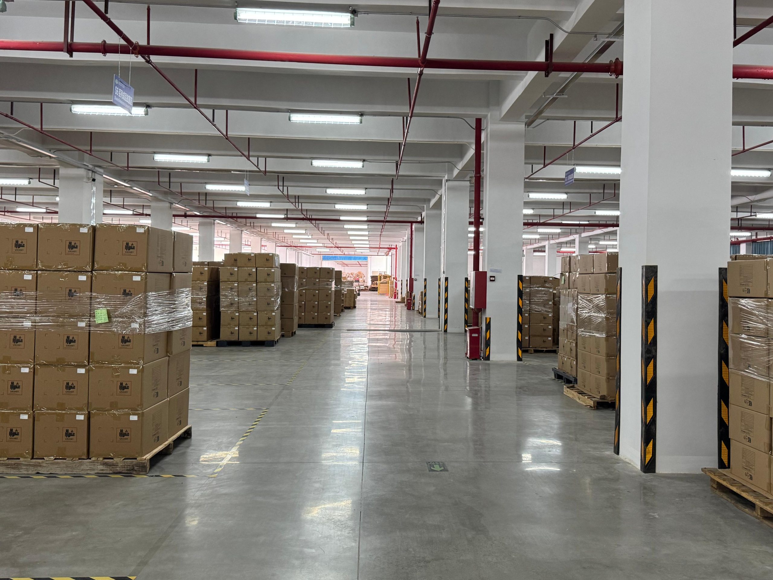 A wide central aisle in an indoor warehouse lined with large blocks of finished product cartons wrapped in stretch film near the pillars.