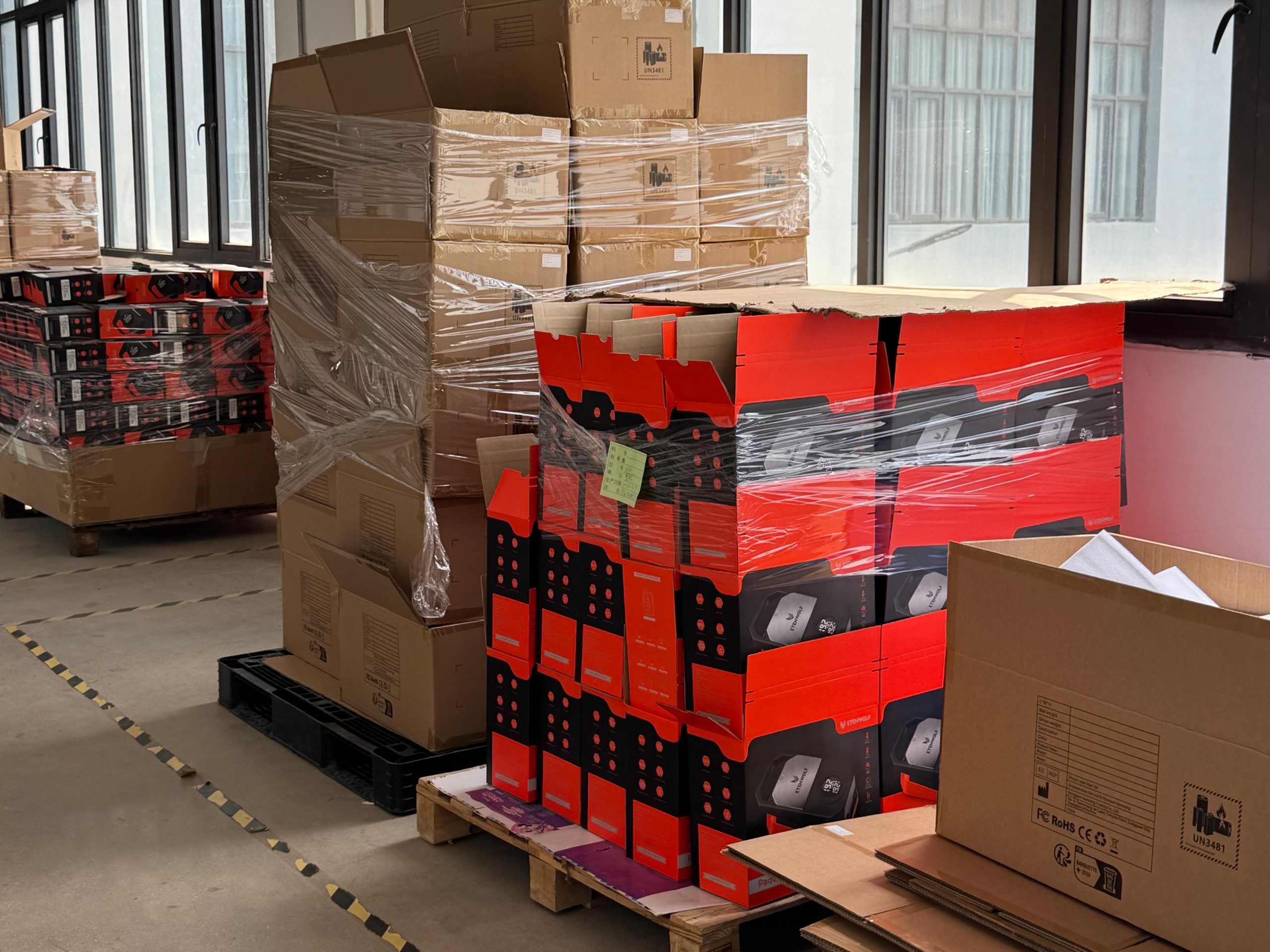 Red and black product color packaging boxes stacked on wooden pallets near warehouse windows, with brown cardboard boxes behind them.