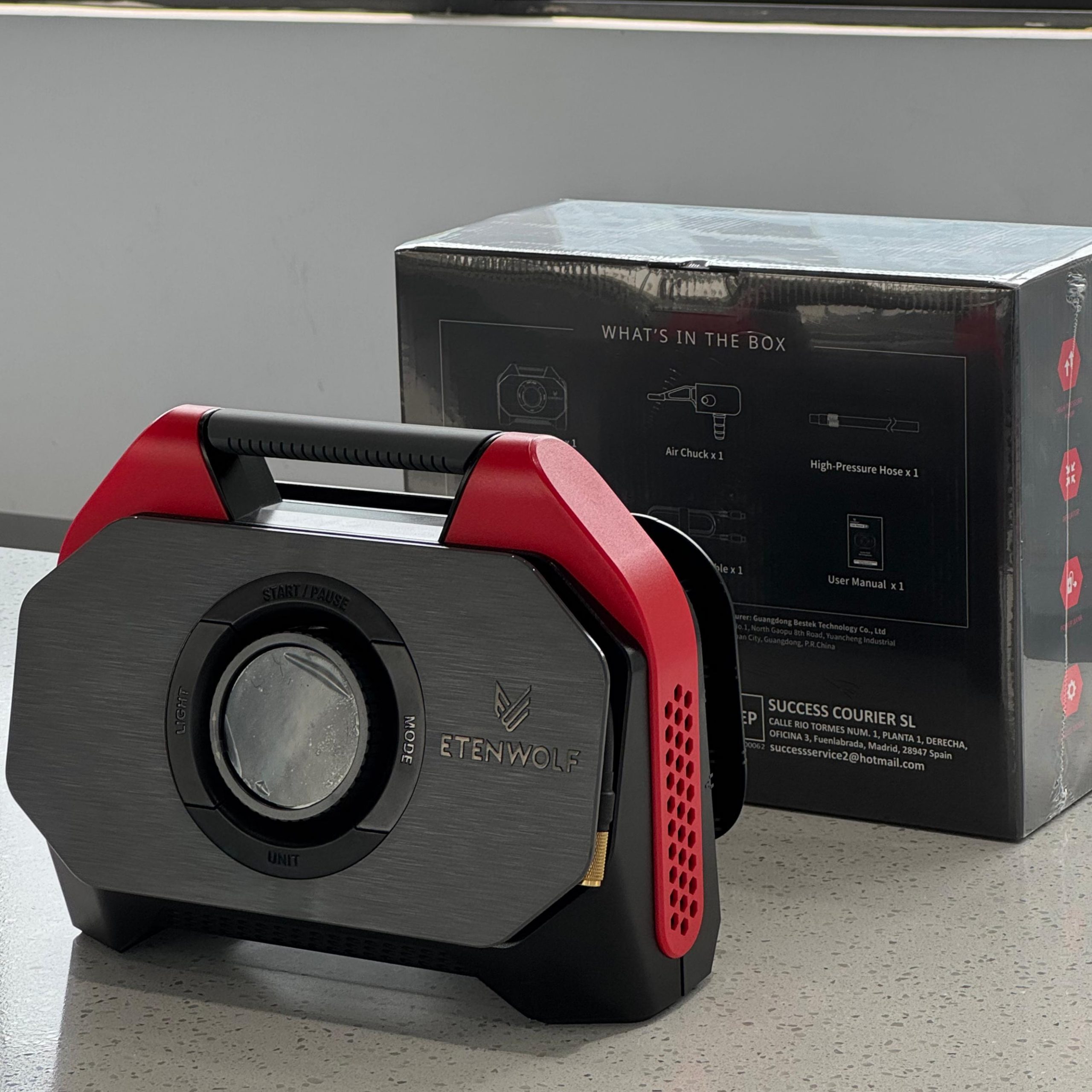 A portable air inflator with a red handle and a grey metallic-textured panel placed next to a black rectangular packaging box on a light marble counter